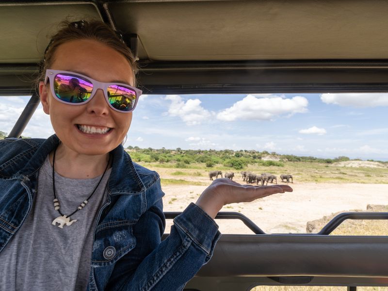 A young woman smiles from the open roof of a safari vehicle, gesturing towards elephants visible through the vehicle's window.