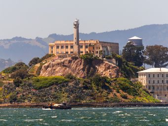 A view of Alcatraz Island from the water. The island is rocky and covered in vegetation, and the former prison is visible on the top of the island. A sailboat is sailing in the foreground, with other boats and ferries in the distance.