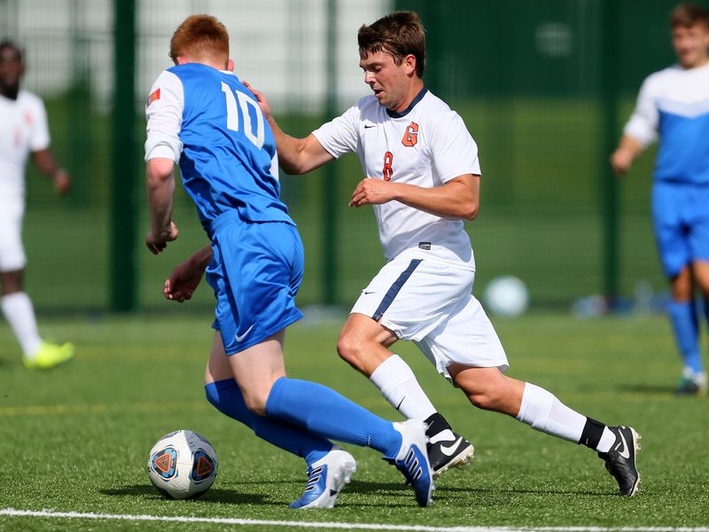 Two young adult soccer players compete for the ball during a game on a green turf field.