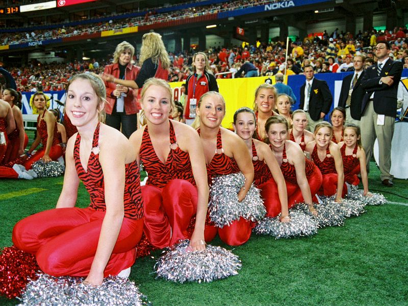 A group of cheerleaders in red and black uniforms pose on the sidelines of a football game.