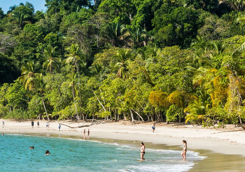 People enjoying a sunny day at Manuel Antonio Beach, with the turquoise ocean lapping at the white sand.