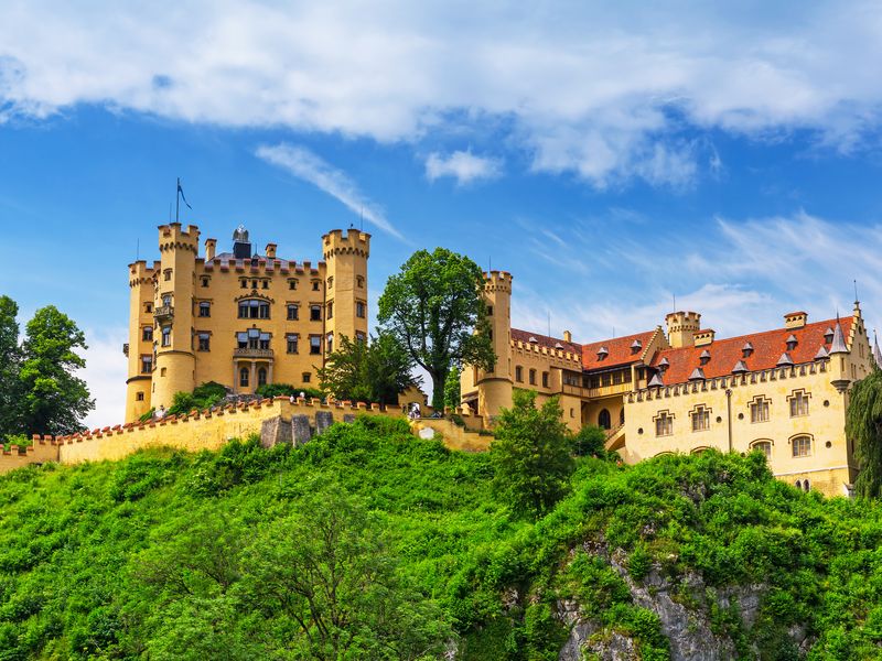 Hohenschwangau Castle in Füssen, Germany. The castle is perched atop of a hill and can be seen in the middle of the image. It is surrounded by lush green grass and trees. The sky is mostly clear and blue.