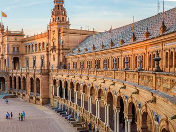 A panoramic view of Plaza de España in Seville, Spain at sunset, with people walking around the square and its central fountain.