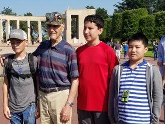 A veteran with his arm around a child and two teenagers, posing together in front of a memorial.