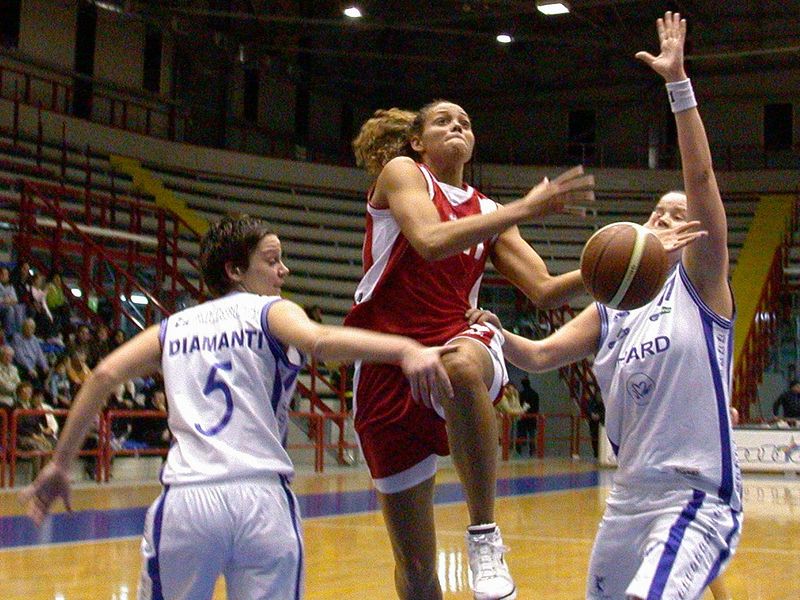 Three women playing basketball in a gym, one jumping to shoot and another defending.