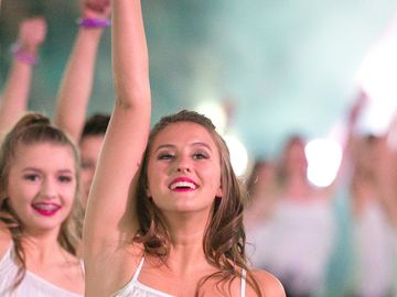 Group of female dancers performing on a field at night with raised arms.