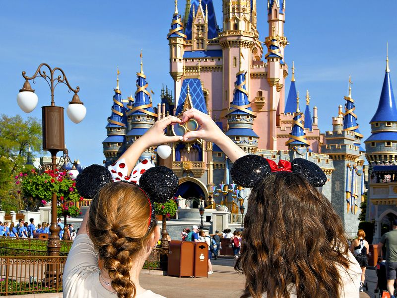 Two teenagers wearing mouse ears stand with their back to the camera and make a heart shape with their hands in front of Cinderella's Castle in Disney World.