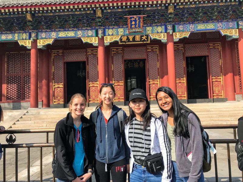 Four teenage girls pose in front of a colorful temple-like building.  The building is red and gold and blue.