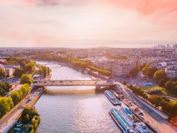 A panoramic aerial view of the Paris cityscape at sunset, showcasing the Eiffel Tower, Seine River, and a vibrant sky.