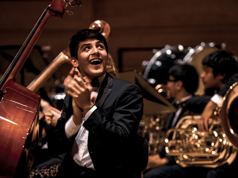 A young adult musician claps and smiles during the curtain call of an orchestra concert. 