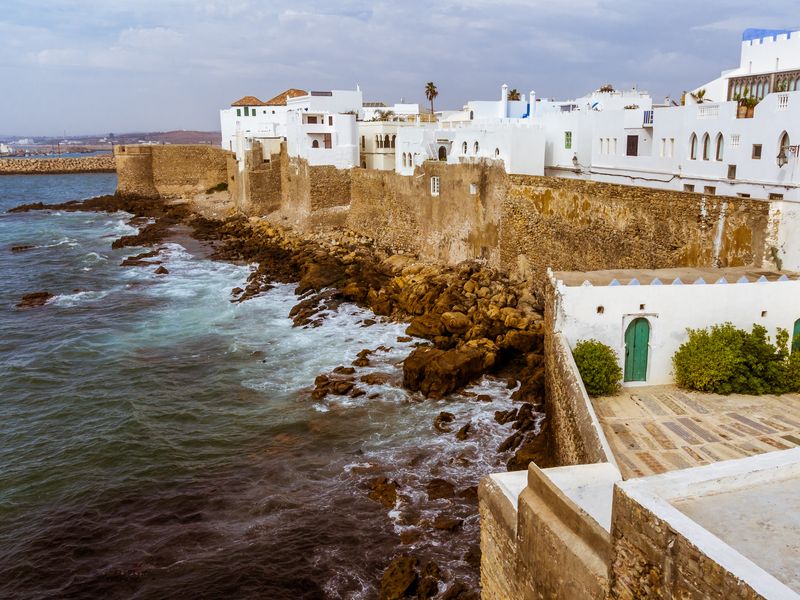 View of Asilah, a coastal town in Morocco with white buildings, brown stone walls, and waves hitting the rocky shore.