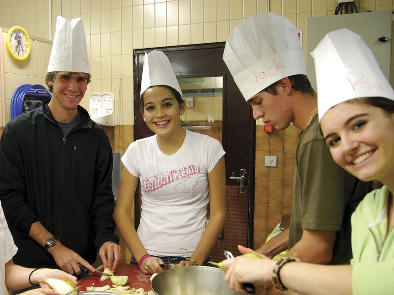 Four young adults in chef hats preparing food in a kitchen.