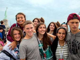 A group of teenagers poses for a photo on a boat in front of the Statue of Liberty.