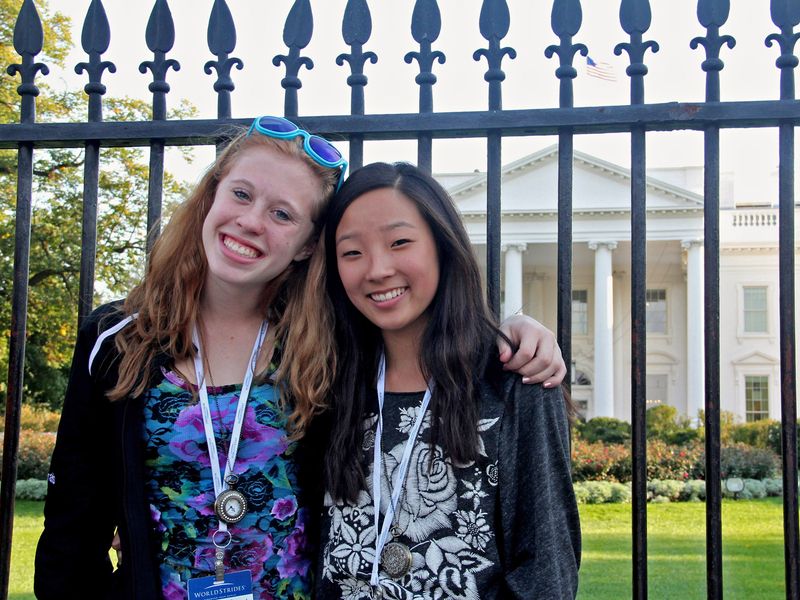 Two teenage girls pose with arms around each other in front of the White House and its fence. The White House is a large white building with columns and several rows of windows. The girls are smiling and both wearing lanyards around their necks.