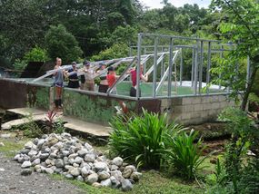 Group of young adults building a metal-framed structure over a concrete base in a tropical area.