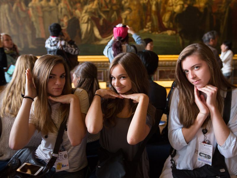 Three young women playfully pose, framing their faces with their hands, in front of a large historical painting in a museum.