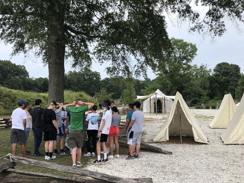 A group of teenagers at a historical reenactment or camp, standing near canvas tents and a large tree.