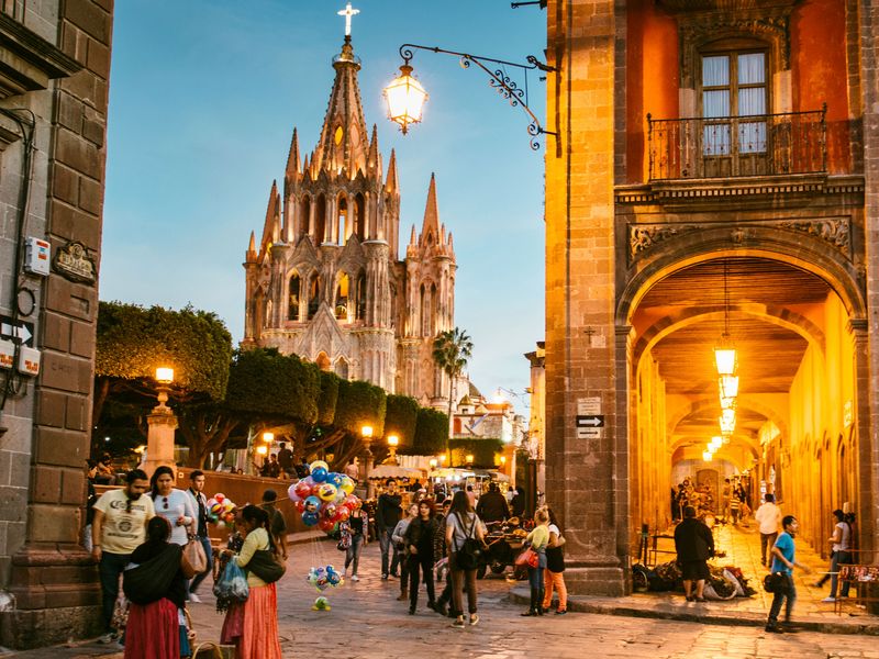 Evening scene in the historic town square of San Miguel de Allende, Mexico, featuring the illuminated Parroquia de San Miguel Arcángel church and a lively crowd.