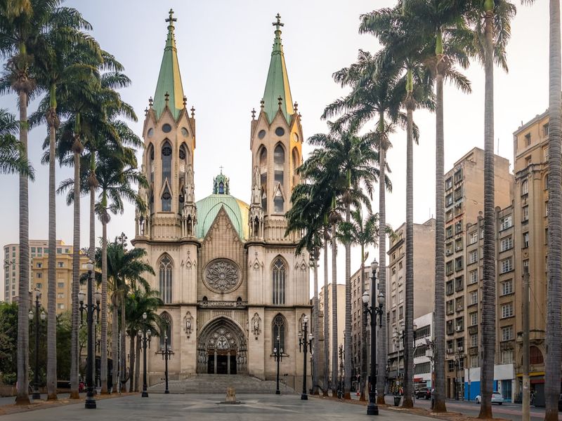 The São Paulo Cathedral in São Paulo, Brazil. It is a large, ornate cathedral with two tall spires and a rose window. Palm trees frame the cathedral, and city buildings are visible in the background.