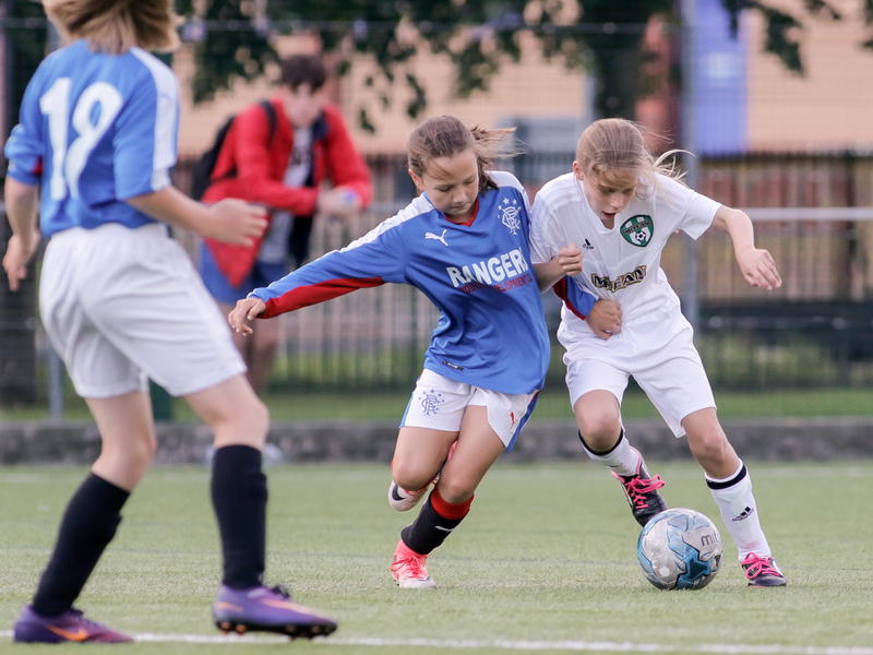 Two young girls playing in a soccer game, one in a blue jersey and the other in white, competing for the ball.