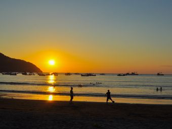 Silhouetted figures play soccer on the beach as the sun sets in a blaze of orange and yellow, casting long shadows on the sand.