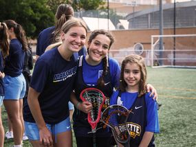 Three smiling girls and women with lacrosse sticks, standing on the field.