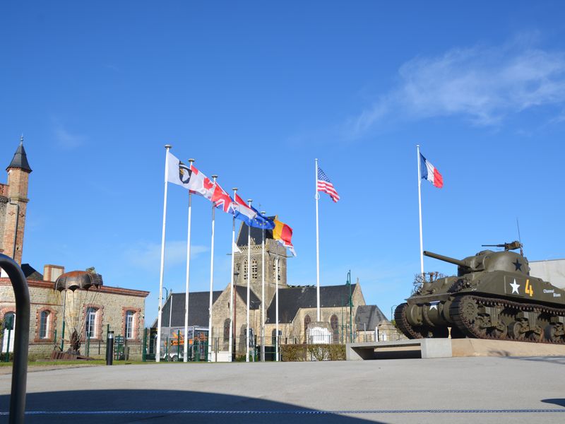 A Sherman tank on display outside in front of a church, with flags from different countries in the background.  This appears to be a World War II memorial or museum in Normandy, France.