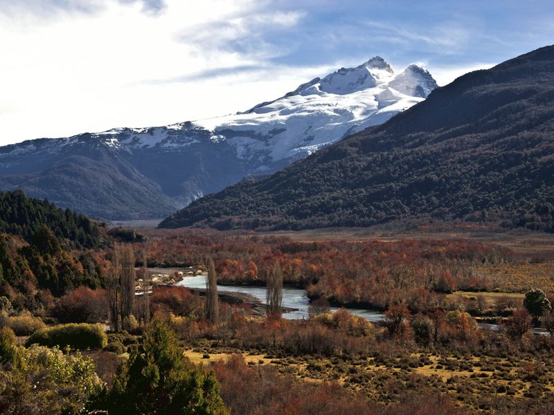 A snow-capped mountain rises above a valley filled with colorful autumn foliage and a winding river in Patagonia.