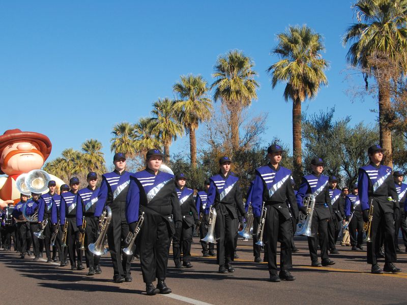 A high school marching band in blue and black uniforms parades down a street lined with palm trees.