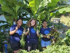 Three young adults proudly display their harvested vegetables in a vibrant community garden.