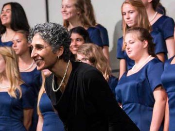 A choir of teenage girls in blue robes performs on stage with a woman conductor.