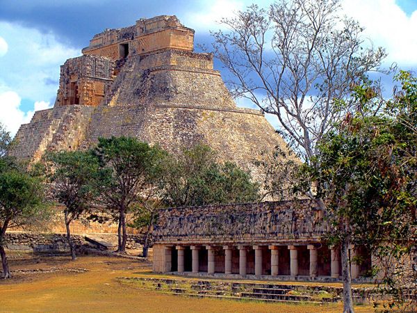 Ancient Mayan Pyramid of the Magician at the Uxmal archaeological site in Yucatan, Mexico. The pyramid stands tall against a backdrop of trees and a cloudy sky. A smaller stone structure with columns is in the foreground.