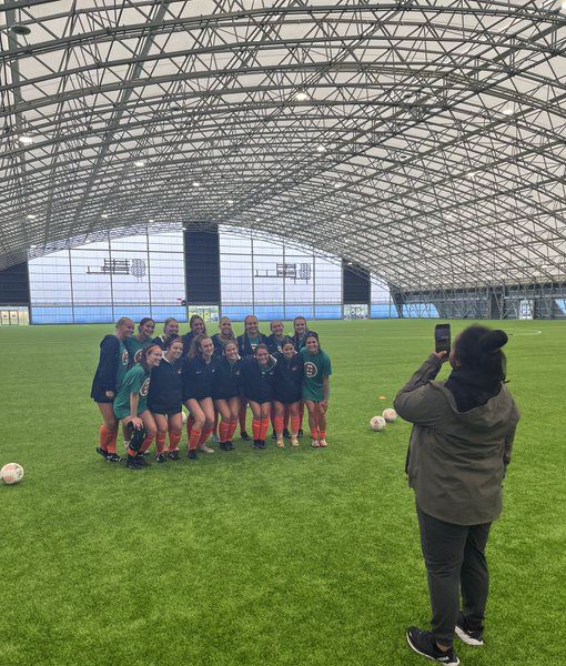 A group of young adult women in soccer uniforms pose for a photo on an indoor soccer field while another young adult woman takes their picture.