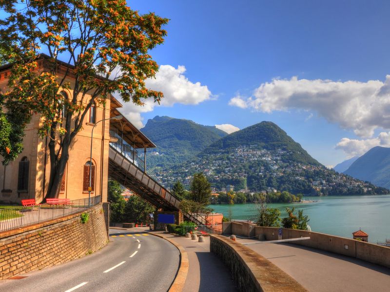 Scenic view of a lakeside town with a building and a winding road in the foreground, and mountains in the background.