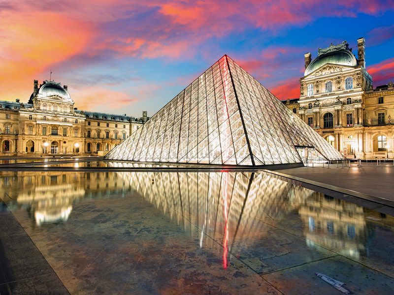 The illuminated Louvre Pyramid at sunset, reflecting in the surrounding water feature, with the historic Louvre Museum buildings in the background in Paris, France.