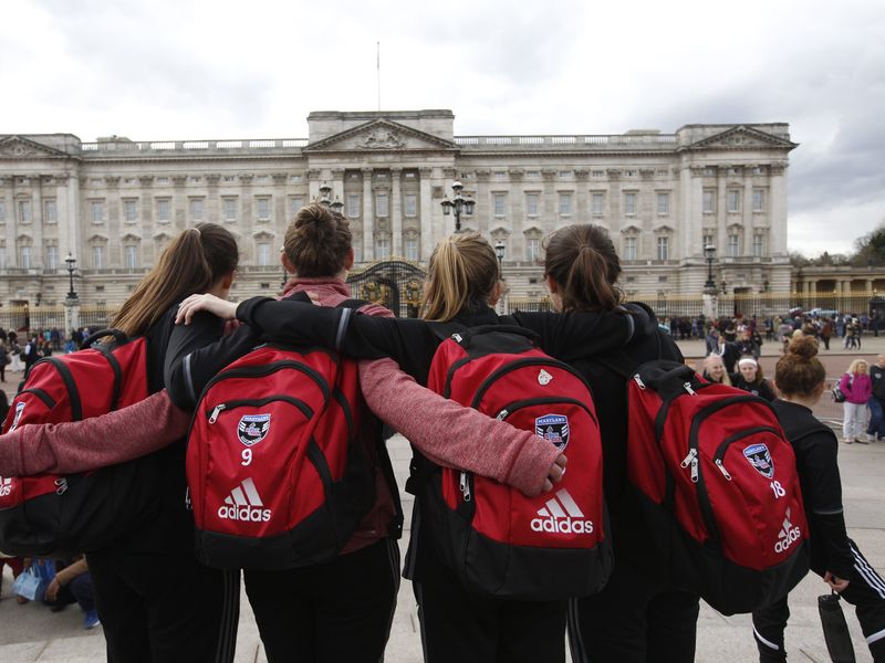 Four young women with red backpacks stand arm in arm, facing away from the camera towards Buckingham Palace.