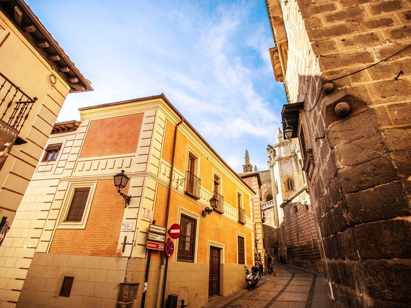 A narrow street in Toledo, Spain, with orange and beige buildings and a cathedral in the background.