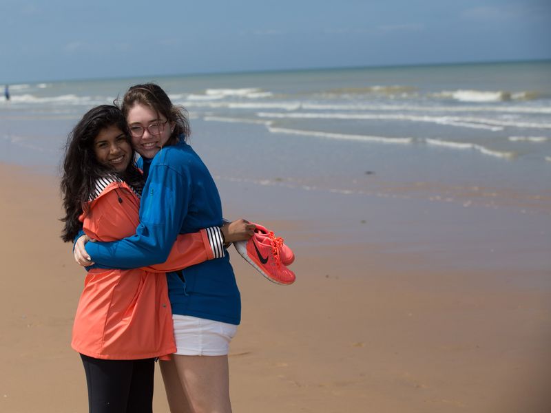 Two young women hug on a beach with ocean waves in the background.