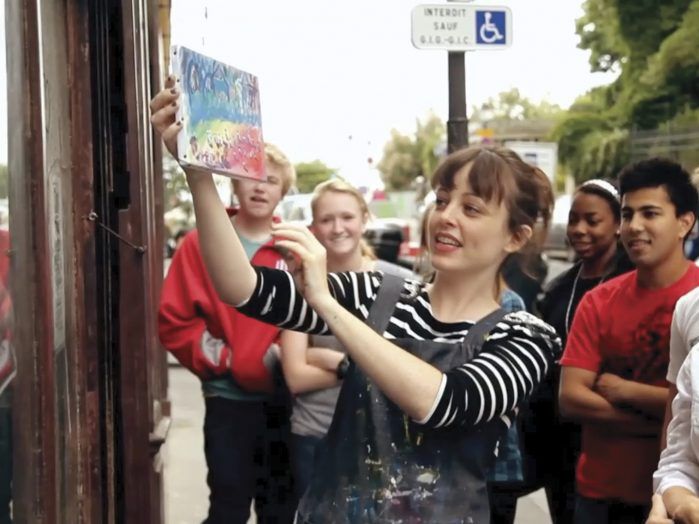 A young woman in a striped shirt and overalls, holding up a colorful painting for a small group of teenagers and young adults to view on a city street.