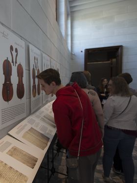 Group of people viewing a museum exhibit about violins and other stringed instruments, including historical descriptions and documents.