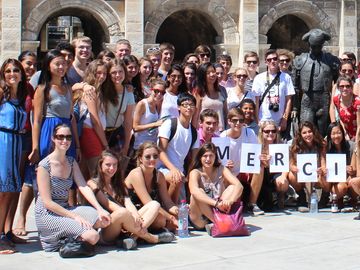 A large group of students holding a "Merci" sign in front of the Arles Amphitheatre.