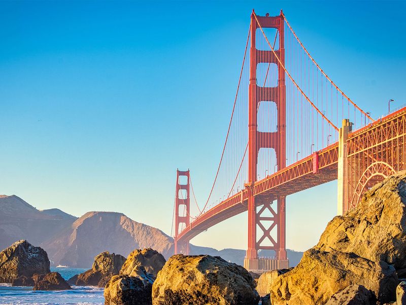 A view of the Golden Gate Bridge from the rocky shoreline, featuring the bridge's red towers and cables against a backdrop of blue sky and water.