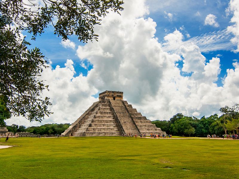 The Chichen Itza pyramid, a well-preserved Mayan ruin in Mexico, stands tall against a vibrant blue sky dotted with fluffy white clouds. Lush green grass surrounds the base of the pyramid, and groups of tourists can be seen exploring the area. The image captures the essence of ancient history, cultural heritage, and the beauty of archaeological wonders.