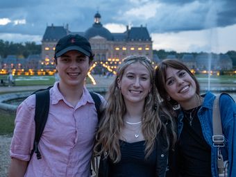 Three young adults posing for a photo in front of a beautifully illuminated chateau at dusk.