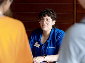 A young adult woman with glasses smiles as she interacts with two people across a table.