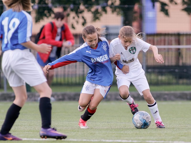 Two young girls playing in a soccer game, one in a blue jersey and the other in white, competing for the ball.