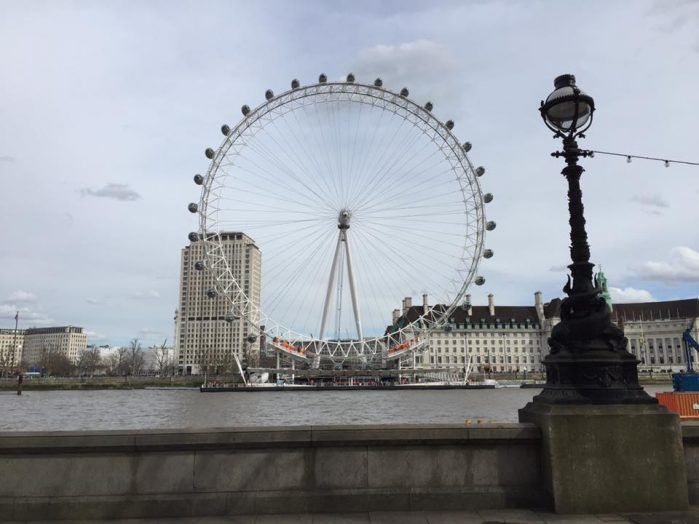 The London Eye Ferris Wheel stands tall against a cloudy sky with the River Thames flowing in the foreground.