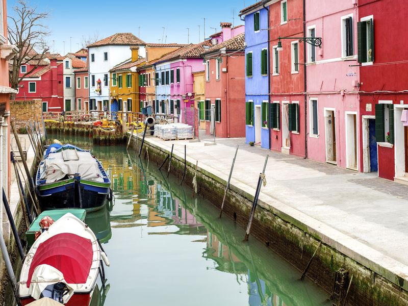 Colorful houses line a canal in Burano, Italy, with boats moored alongside. The water reflects the vibrant colors of the buildings.