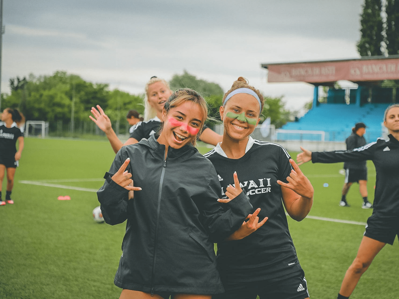 Two female soccer players giving the hang loose sign on a field.