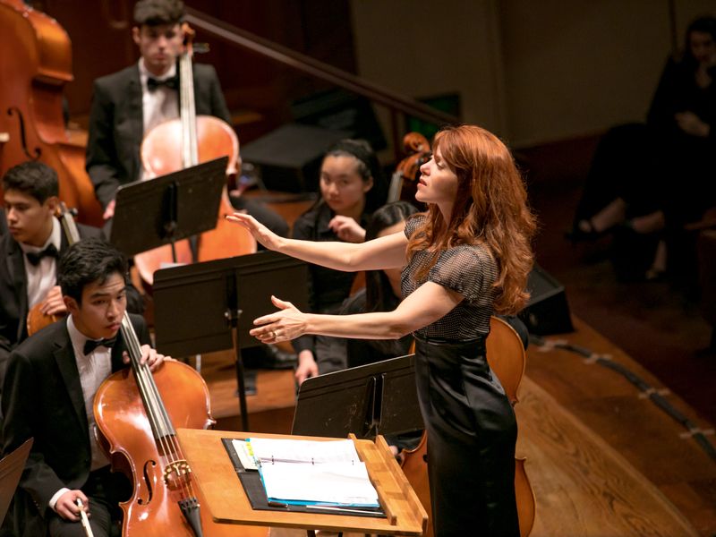 A woman in a black dress conducts a youth orchestra on stage.